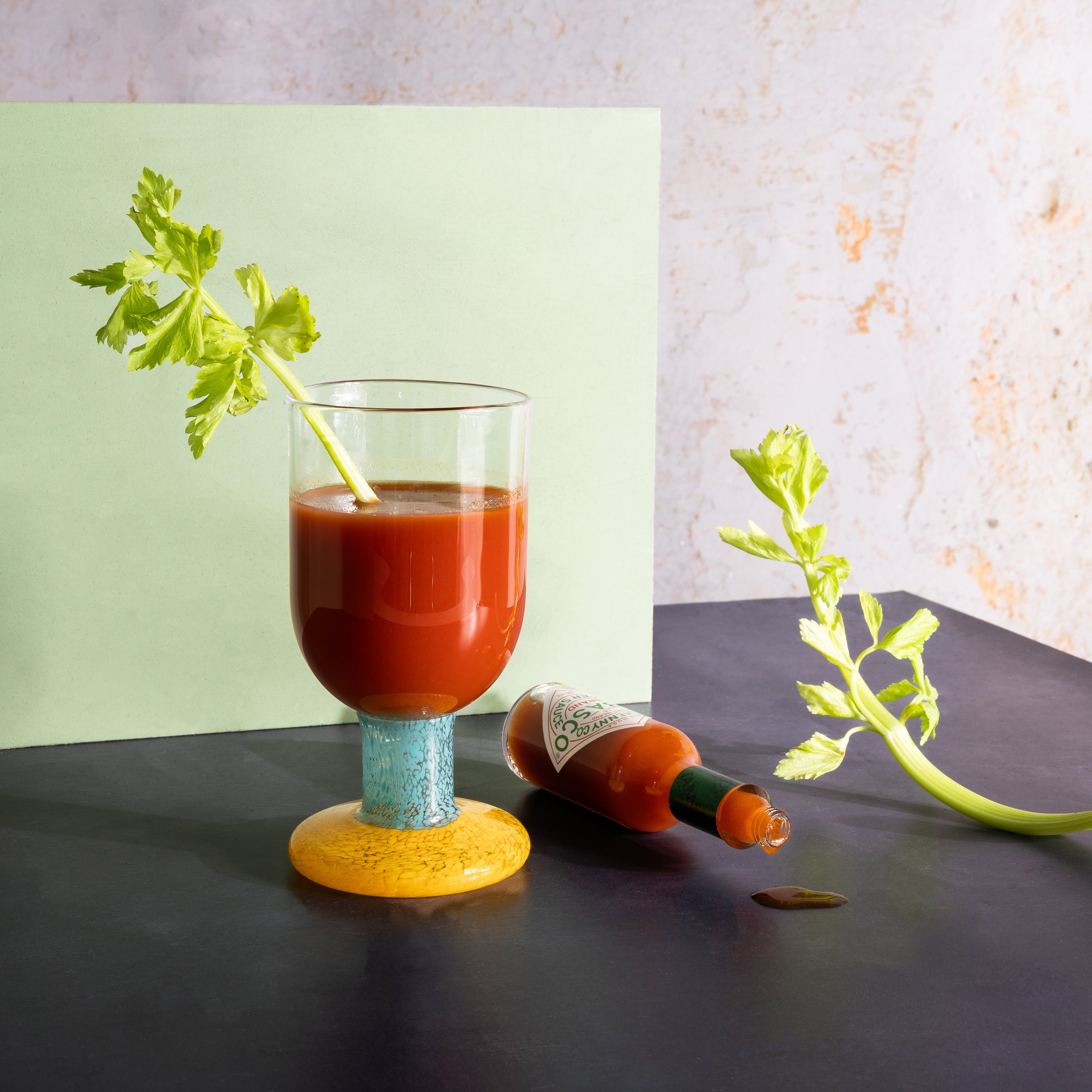 Glass of red juice with celery stick on a dark surface, bottle of hot sauce nearby.